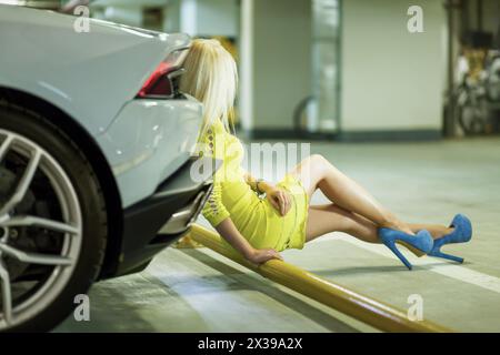 Junge Frau in gelbem Kleid sitzt auf gelbem Rohr und lehnt sich zurück auf den Kofferraum eines modernen weißen Autos in der Tiefgarage. Stockfoto