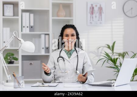 Eine professionelle hispanische Frau, die als Ärztin arbeitet, lächelt, während sie in einem gut organisierten Klinikbüro Gesundheitslösungen bespricht und Vertrauen und Fachwissen ausstrahlt. Stockfoto