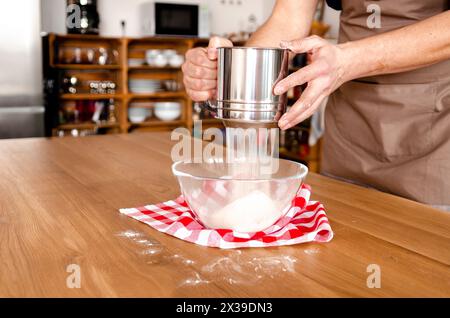 Die Person siebt Mehl in eine Schüssel auf einem Holztisch, um das Rezept vorzubereiten Stockfoto