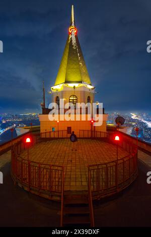 Der Mann steht auf einer runden Aussichtsplattform mit roten Lichtern auf dem Dach eines hohen Gebäudes in der Nachtstadt Stockfoto