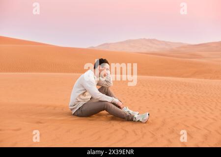 Junger Mann in Jeans sitzt auf gelbem Sand in der Wüste an sonnigen heißen Tagen Stockfoto