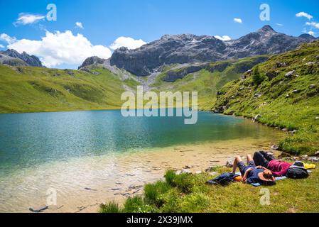 Paare (nicht erkennbare Personen), die sich am Ufer des wunderschönen La Plagne Sees in den französischen Alpen entspannen. Andere Wanderer gehen den Hügel hinauf. Savoie, Frankreich Stockfoto