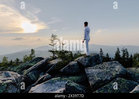 Ein Mann steht selbstbewusst auf dem Gipfel eines felsigen Berges mit Blick auf eine riesige Landschaft darunter. Er wirkt klein auf dem zerklüfteten Gelände, Showcasin Stockfoto
