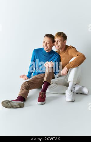 Ein liebevolles schwules Paar in legeren Gewändern, das eng zusammen vor grauem Hintergrund sitzt. Stockfoto