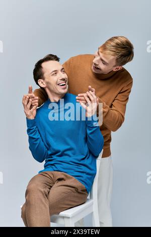 Zwei Männer in lässiger Kleidung sitzen auf einem weißen Stuhl, teilen sich Lachen und einen besonderen Moment. Stockfoto
