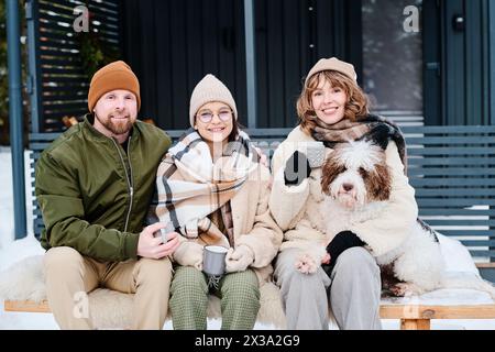 Porträt einer glücklichen kaukasischen Familie von drei Personen mit lagotto romagnolo Hund, der draußen auf einer Holzbank sitzt, an kalten Wintertagen Stockfoto