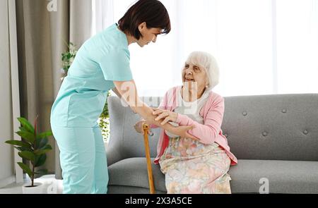 Die Krankenschwester übergibt den Stock an eine ältere Frau und hilft ihr, sich in Einem Pflegeheim im Raum zu bewegen Stockfoto