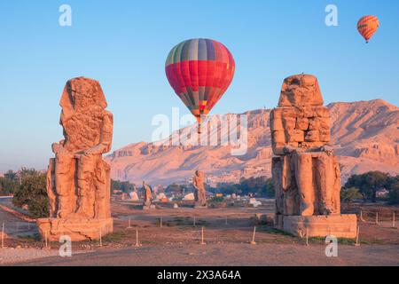 Farbenfrohe Heißluftballons fliegen über die Memnonkolosse bei Sonnenaufgang am Westufer von Luxor, Ägypten Stockfoto