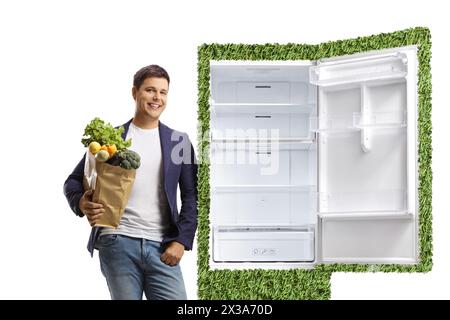 Junger Mann, der eine Einkaufstasche hält und sich auf einen grünen umweltfreundlichen Kühlschrank gestützt hat, der auf weißem Hintergrund isoliert ist Stockfoto