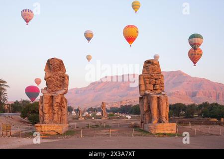 Farbenfrohe Heißluftballons fliegen über die Memnonkolosse bei Sonnenaufgang am Westufer von Luxor, Ägypten Stockfoto