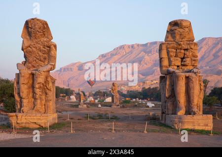 Ein farbenfroher Heißluftballon, der bei Sonnenaufgang über die Memnonkolosse am Westufer von Luxor, Ägypten, fliegt Stockfoto