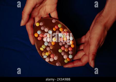 Großes Schokoladen-Osterei mit bunten Bonbons in weiblichen Händen Dunkle Sicht von der Tischplatte. Osterspezialitäten und Süßigkeiten. Festliche Essensgeschichte. Eierjagd Stockfoto