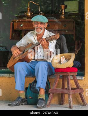 Ein älterer Mann saß auf einer Fensterbank vor einem Geschäft und spielte eine Gitarre für Touristen, um in der Altstadt von Havanna, Kuba, zu betteln. Stockfoto