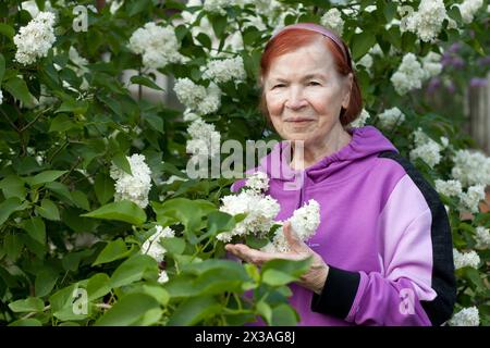 Außenporträt einer alten Seniorin. Schöne ältere Frau lächelt vor dem Hintergrund blühender weißer Flieder im Frühlingspark. Stockfoto