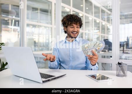 Fröhlicher junger Mann in einem hellblauen Hemd, der fröhlich einen Stapel Geld an seinem Schreibtisch hält und zählt, mit Laptop und Tablet Geschenk. Stockfoto