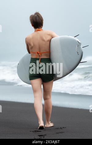 Rückansicht einer nicht erkennbaren Surferin, die am Sandstrand mit Surfbrett in der Hand läuft. Der Hintergrund ist mit beruhigendem Rauschen der Meereswellen gefüllt Stockfoto