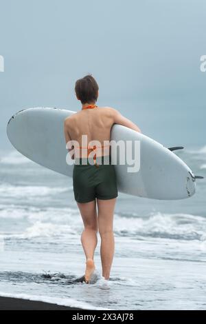 Rückansicht einer Surferin am Sommerstrand, die ihr Board trägt und wie eine unaufhaltsame Kraft aussieht, während sie entlang des Sandes schreitet Stockfoto