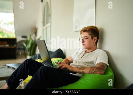 Transgender Pro arbeitet auf einem Laptop aus einem gemütlichen Sitzsack in einem modernen Coworking Space. Die Person konzentriert sich auf den Bildschirm und genießt ein flexibles Arbeitsumfeld. Inklusive Stockfoto