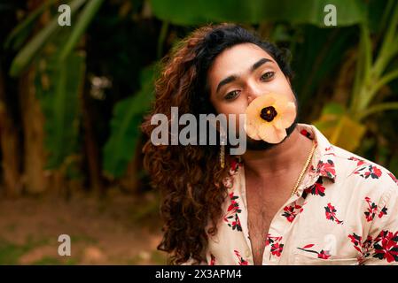 Schwarzer schwuler Mann mit Blume im Mund posiert in der Natur. Stilvoller Mann mit lockigen Haaren, Blumenhemd, goldene Halskette. LGBTQ-Stolz, Ausdruck der Identität Stockfoto