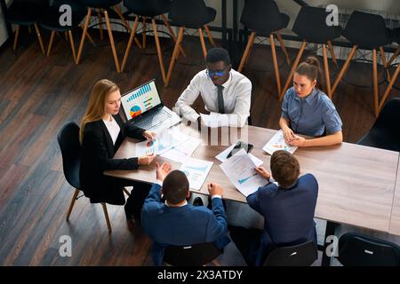Multirassisches Team am Verhandlungstisch des Büros, geführt von weiblicher CEO. Geschäftsfrau analysiert Diagramm auf Laptop, Mitarbeiter diskutieren Strategie. Unternehmenstreffen Stockfoto