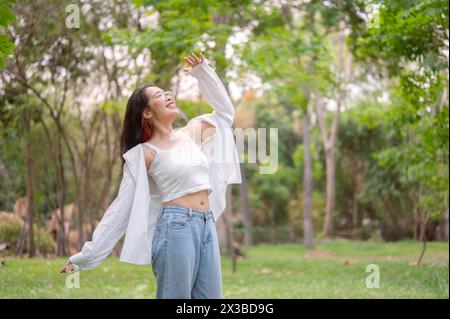 Eine junge, glückliche asiatische Frau in Brille, mit weißem Oberteil und Jeans, tanzt freudig in einem üppig grünen Park, drückt Freiheit aus und genießt die Ruhe Stockfoto