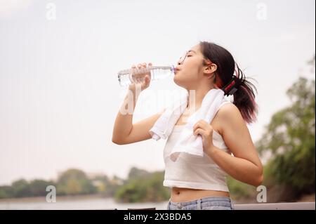 Eine junge asiatische Frau in Gläsern und einem weißen Tanktop trinkt aus einer Wasserflasche, während sie ein Handtuch um den Hals hält und sich nach einem Spaziergang im Freien abkühlt Stockfoto