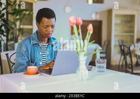 Frau, die einen Laptop mit Kaffeetasse und Tulpen auf einem Tisch im sonnendurchfluteten Restaurant benutzte Stockfoto