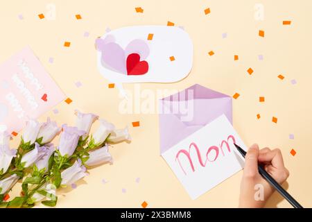 Handmodell mit schwarzem Stift, auf dem das Wort „Mama“ auf einer Karte steht. Ein Blumenstrauß mit viel Konfetti vor beigefarbenem Hintergrund. Internatio Stockfoto