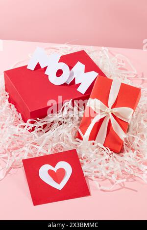 Drei Buchstaben bilden zusammen das Wort Mom, das auf einem roten Podium in rechteckiger Form steht. Eine Geschenkbox und eine Karte. Ein Stapel Kraftpapier dekoriert für Stockfoto
