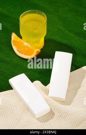 Minimale Szene von zwei Flaschen Sonnencreme oder Feuchtigkeitscreme, die auf dem Schal verziert sind. Grünes Gras enthielt eine Tasse Orangensaft und eine Orangenscheibe. Conta Stockfoto