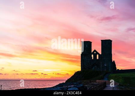 Der Morgenhimmel über den Ruinen der Doppeltürme der angelsächsischen Kirche bei Reculver an der Küste von Kent in der Nähe der Herne Bay. Die Türme haben fast eine Silhouette vor einem leuchtend gelben und rotrosa Himmel. Stockfoto