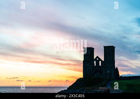 Der Morgenhimmel über den Ruinen der Doppeltürme der angelsächsischen Kirche bei Reculver an der Küste von Kent in der Nähe der Herne Bay. Die Türme haben fast eine Silhouette vor einem leuchtend gelben und rotrosa Himmel. Stockfoto