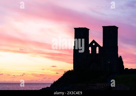 Der Morgenhimmel über den Ruinen der Doppeltürme der angelsächsischen Kirche bei Reculver an der Küste von Kent in der Nähe der Herne Bay. Die Türme haben fast eine Silhouette vor einem leuchtend gelben und rotrosa Himmel. Stockfoto