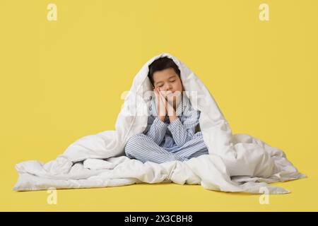 Verschlafener kleiner asiatischer Junge im Pyjama mit Decke auf gelbem Hintergrund Stockfoto