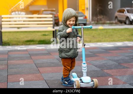 Der süße, glückliche Kleinkind, der drei Jahre alt ist, steht in der Nähe eines Rollers auf dem Spielplatz in einer Wohnanlage. Aktive Frühlingsaktivität für Kinder. Kind Stockfoto