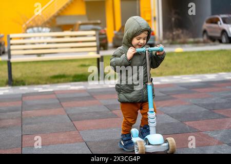 Der süße, glückliche Kleinkind, der drei Jahre alt ist, steht in der Nähe eines Rollers auf dem Spielplatz in einer Wohnanlage. Aktive Frühlingsaktivität für Kinder. Kind Stockfoto