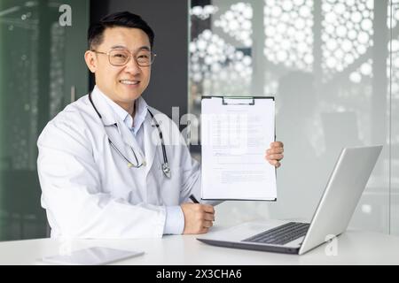 Ein reifer, asiatischer Arzt lächelt warm, während er ein Klemmbrett hält und in einem gut beleuchteten, modernen Klinikbüro neben einem Laptop sitzt. Stockfoto