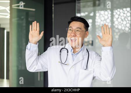 Ein freudiger reifer asiatischer Arzt feiert mit Tanz in seiner Klinik, trägt ein Stethoskop und einen Labormantel und zeigt Glück und Positivität in der Gesundheitsversorgung. Stockfoto