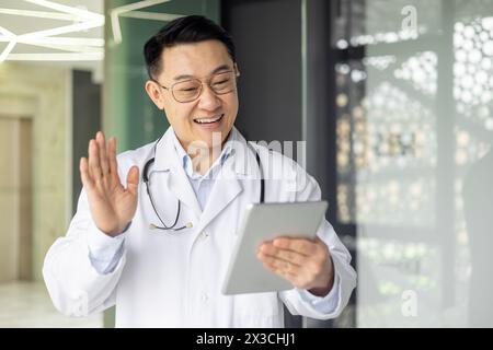 Freundlicher asiatischer reifer männlicher Arzt, der mit einem Tablet einen Videoanruf führt. Er lächelt und winkt, positioniert in einer modernen Klinik. Stockfoto