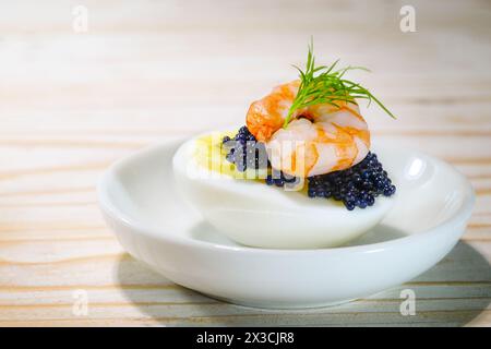 Halbierte gekochte Eier mit schwarzem Kaviar, Garnelen und Dill garnieren auf einem kleinen weißen Teller und einem Holztisch, Fingerfood Snack für Feiertage wie Ostern, CH Stockfoto