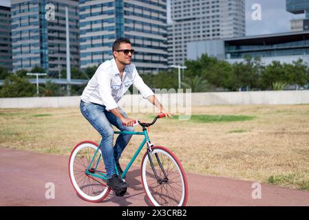Modischer junger Mann, der in einem Stadtpark mit einem Fixie-Fahrrad fährt, mit modernen Gebäuden im Hintergrund, die ein Beispiel für umweltfreundliches Pendeln darstellen Stockfoto