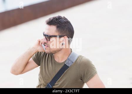 Stilvoller Mann in Sonnenbrille und Kuriertasche, der telefoniert und sich in urbaner Umgebung auf ein Oldtimer-Fahrrad stützt, was auf Mobilität und Stadtleben hindeutet Stockfoto