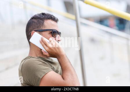 Stilvoller Mann in Sonnenbrille und Kuriertasche, der telefoniert und sich in urbaner Umgebung auf ein Oldtimer-Fahrrad stützt, was auf Mobilität und Stadtleben hindeutet Stockfoto