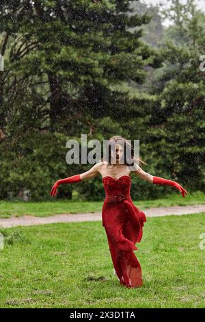 Eine junge Frau in einem auffälligen roten Kleid und langen Handschuhen läuft anmutig im Regen und nimmt die natürlichen Elemente um sie herum auf. Stockfoto