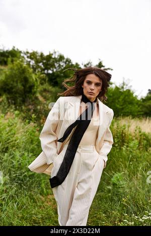 Eine schöne junge Frau in weißem Anzug und schwarzer Krawatte steht selbstbewusst auf einem Feld und spürt die Sommerbrise. Stockfoto