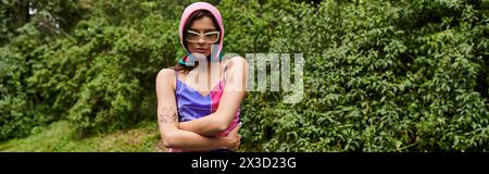 Eine wunderschöne junge Frau in einem leuchtenden lila-blauen Kleid steht anmutig vor üppigen grünen Bäumen und genießt die Sommerbrise. Stockfoto