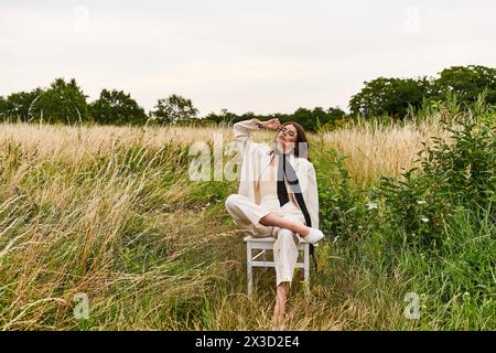 Eine junge Frau, in weiß gekleidet, sitzt friedlich auf einem Stuhl inmitten eines üppigen Feldes und taucht in die Wärme der Sommerbrise ein. Stockfoto