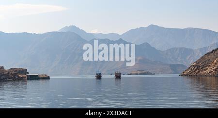 Zwei Boote segeln sanft durch das ruhige Wasser der Fjorde von Khasab, umgeben von der majestätischen Umarmung zerklüfteter Berge, und enthüllen eine Reise, wo Sie hinkommen Stockfoto