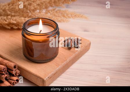 Massage duftende brennende Kerze in bernsteinfarbenem Glas, Winter Hygge Inneneinrichtung auf Holztablett mit Zimtstäbchen und Anis, Kopierraum. Sojawachs Stockfoto