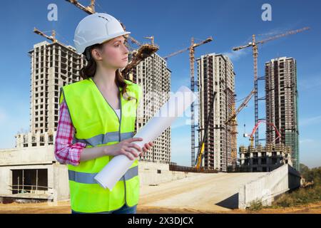 Die Arbeiterin steht mit Zeichnungen auf dem Hintergrund mit vielen hohen Gebäuden im Bau und Kränen unter blauem Himmel, сollage Stockfoto